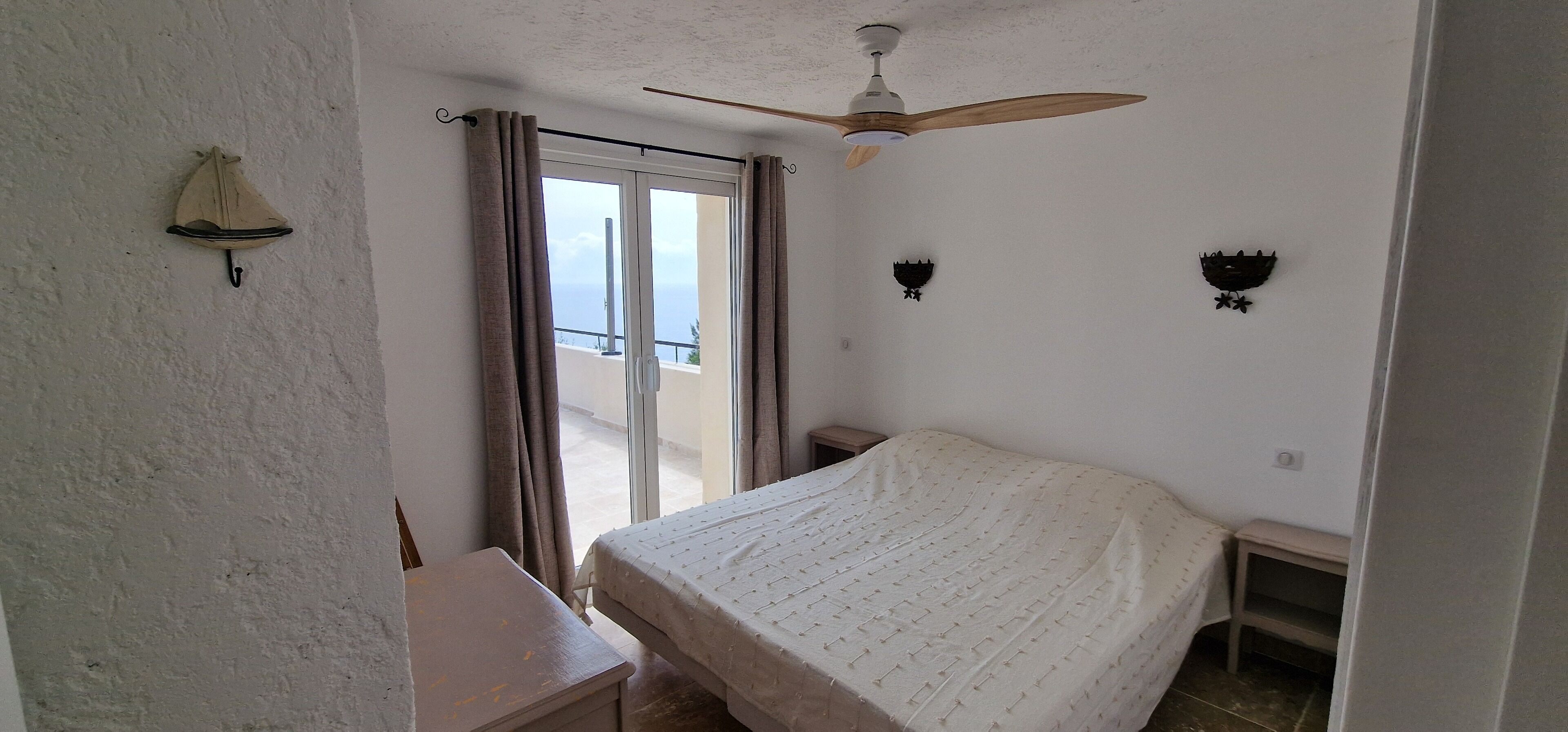 Ein Schlafzimmer mit Bett, Deckenventilator, Fenster mit Vorhängen und Blick aufs Meer.