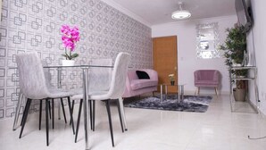 Een moderne woonkamer met een glazen eettafel, twee grijze stoelen, een roze bank, een klein bijzettafeltje en een decoratieve plant.
