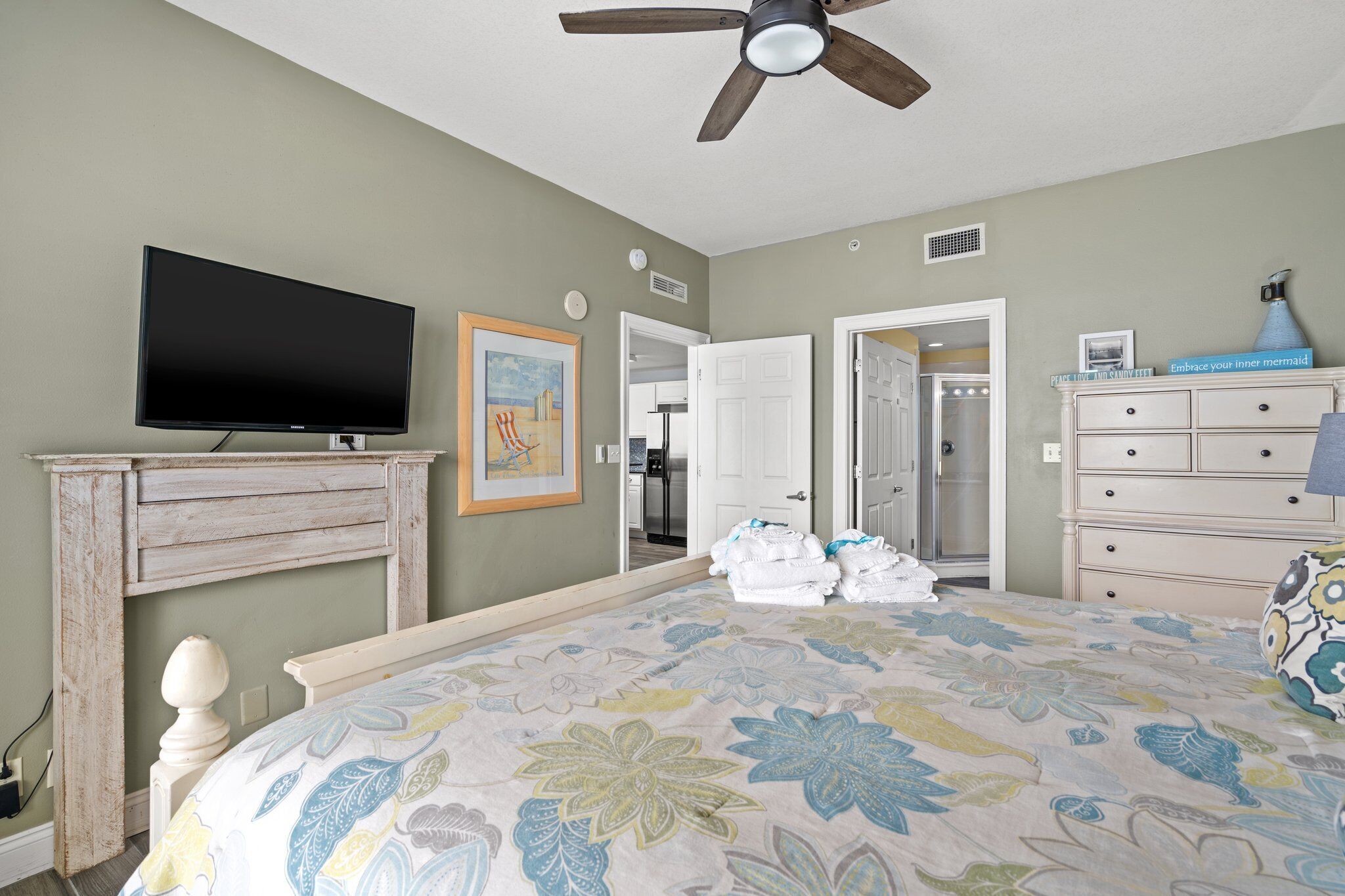 A bedroom with a bed, a television, a ceiling fan, a dresser, and a door leading to a bathroom.