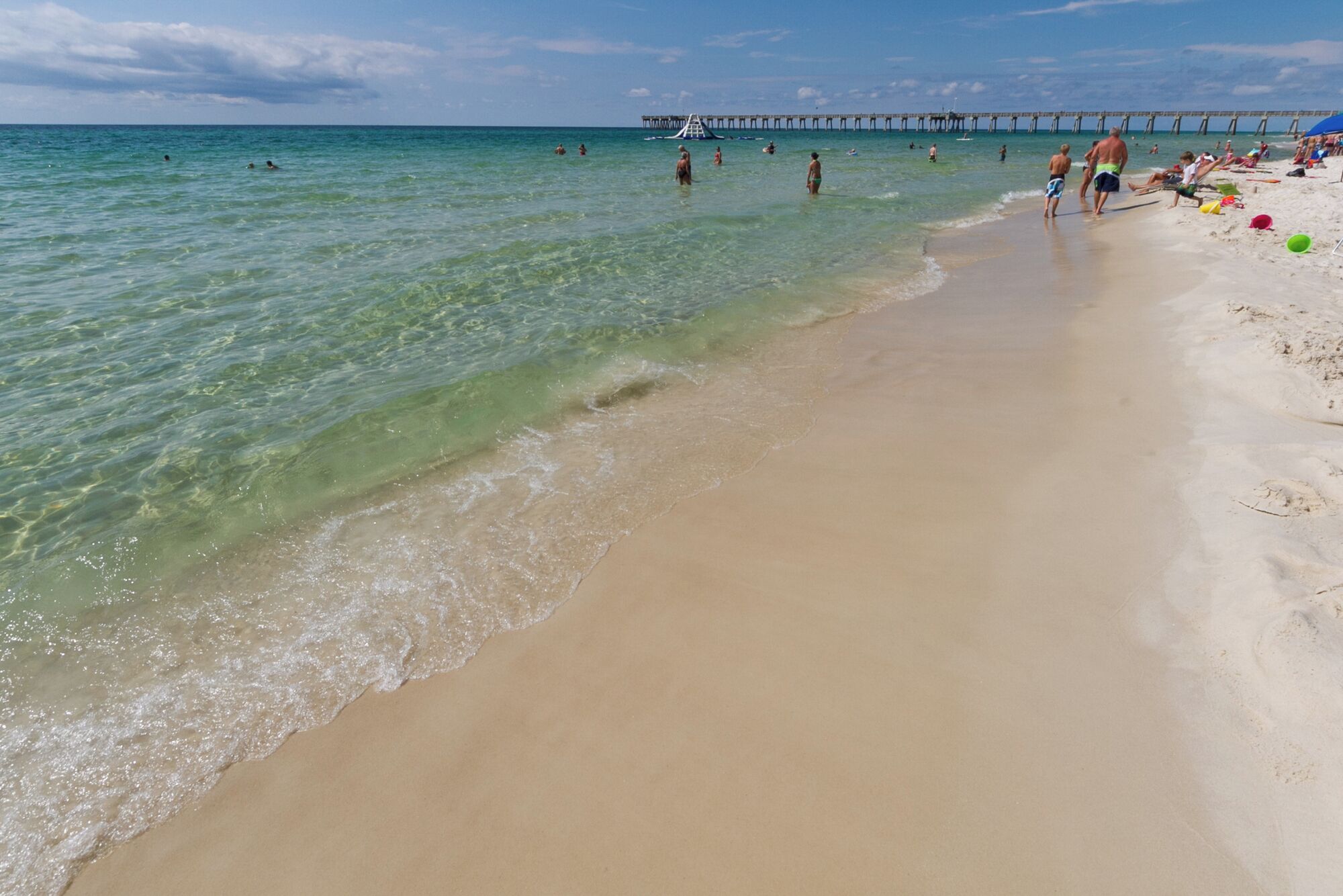 A beach with clear water, a pier in the background, and people swimming and walking on the sand.