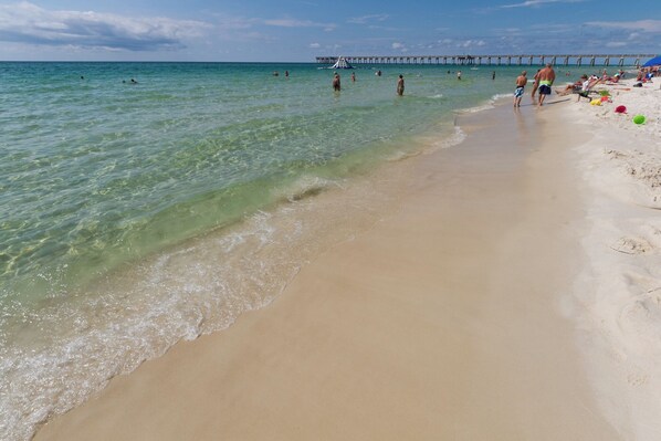 A beach with clear water, a pier in the background, and people swimming and walking on the sand.