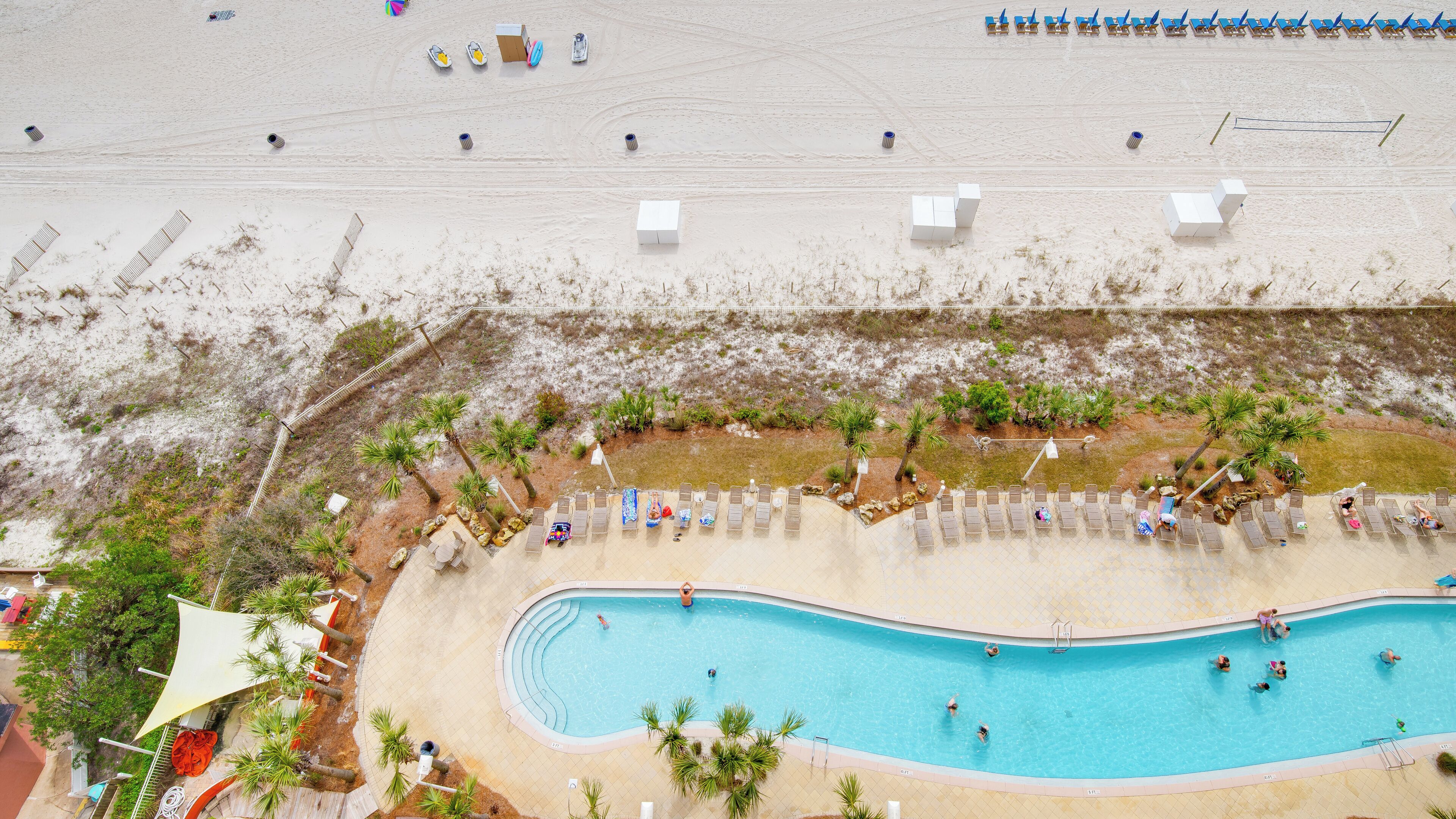 An aerial view of a resort with a swimming pool, beach chairs, and a sandy area.