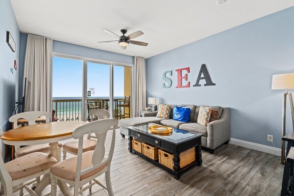 A living room with a view of the sea, a ceiling fan, a sofa, a coffee table, and a dining table with chairs. - Calypso By Blue Swell (Panama City Beach)