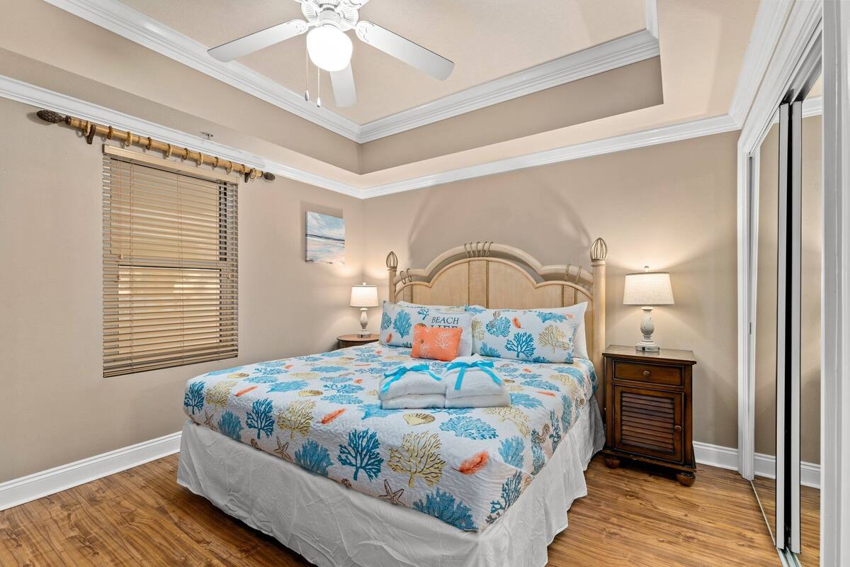 A bedroom with a large bed, a ceiling fan, a nightstand with a lamp, and a window with blinds.