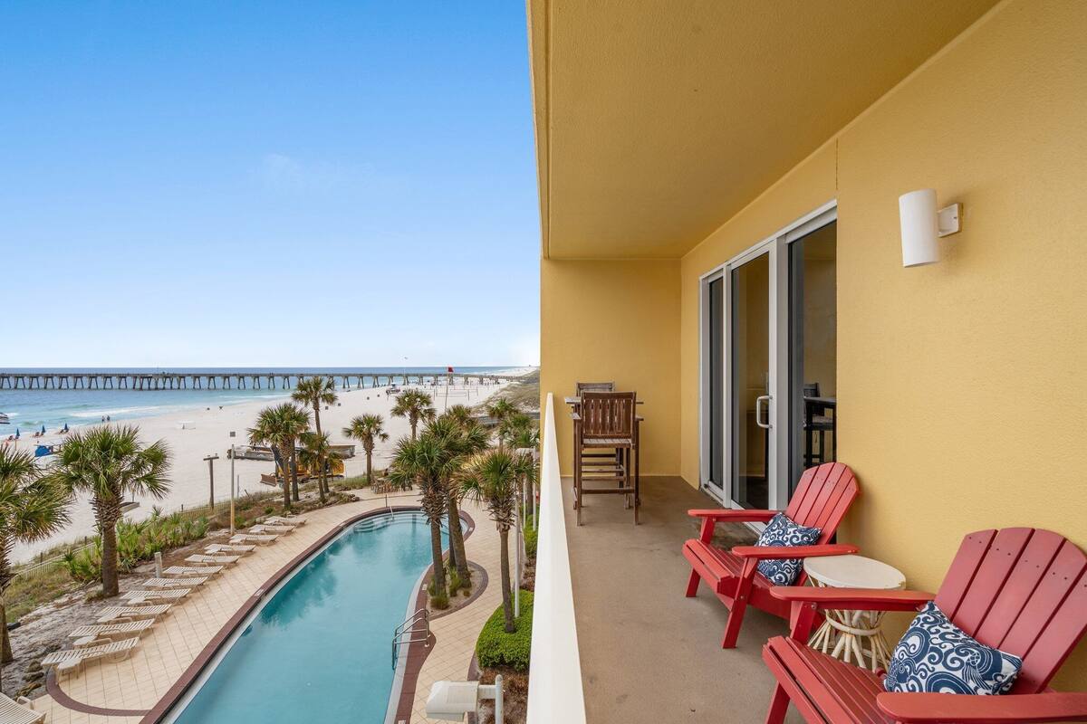 A pool with a view of the beach and a pier, and a balcony with red chairs and a table.