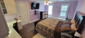 A bedroom with a bed, a TV, a ceiling fan, and a window with blinds.