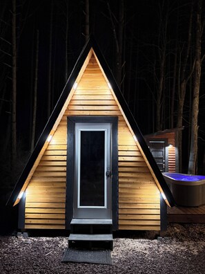 A small, triangular wooden cabin with a dark door, illuminated by lights around the entrance. - Glamp in this Cozy Tiny A-Frame Cabin: Electricity, Heating, AC, Wi-Fi & Hot Tub (Newfoundland)