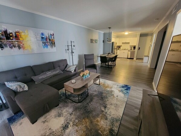 Un salon moderne avec un canapé d’angle, une table basse et un tapis. On y trouve également un coin repas avec une table et des chaises. La cuisine est visible au fond.