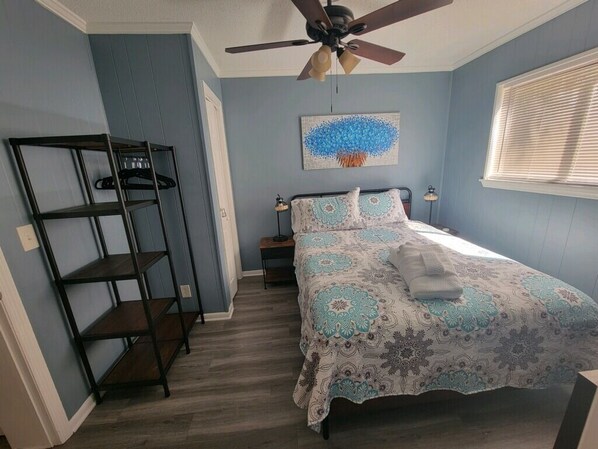 Un dormitorio con cama, ventilador de techo, estante en la pared y ventana con persianas.