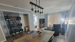 Un comedor con mesa de madera, sillas y una araña. Se observa un televisor al fondo y una estantería con diversos objetos.