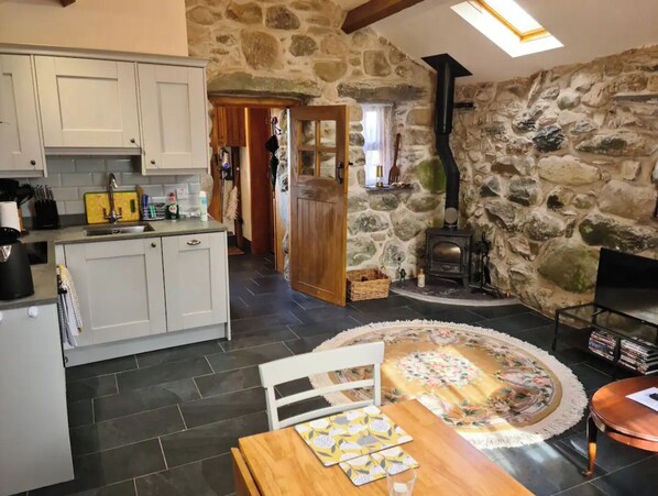 Une cuisine rustique aux murs de pierre, dotée d’une cheminée et d’un coin repas agrémenté d’une nappe à motifs floraux.