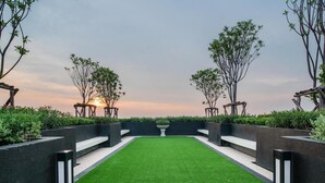 A landscaped garden with a central lawn, surrounded by low hedges and trees, and a sunset in the background. - Lovely 2-bed Apart - 19th Floor Notting Hill Bldg (Samut Prakan)