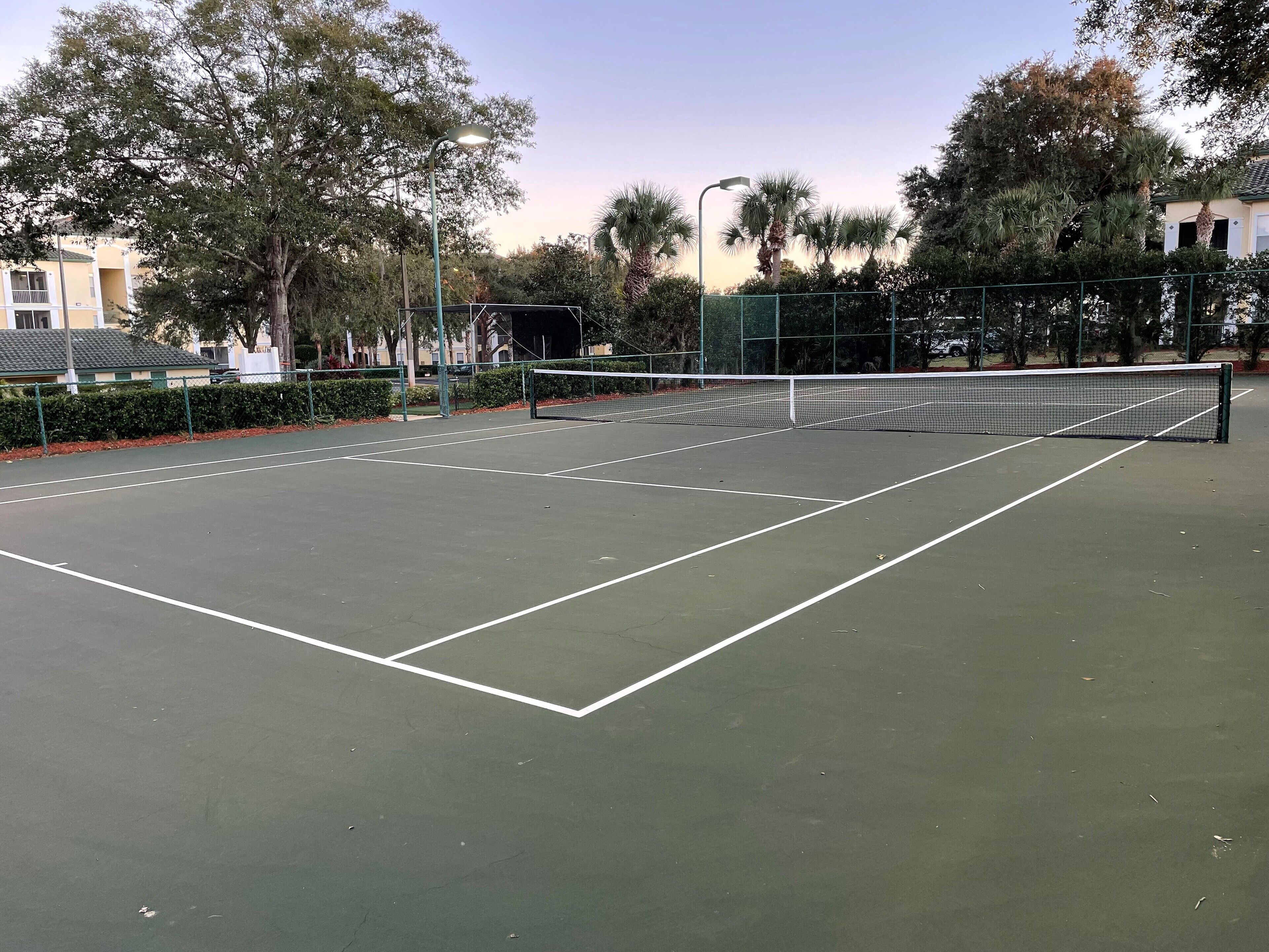 An outdoor tennis court with a net, surrounded by trees and a fence.