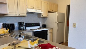 A kitchen with light wooden cabinets, a white stove, a white refrigerator, and a sink with a woven mat and a red cloth on the counter. - Lovely Condo Stay Disney Orlando, 2Bed/2Bath (Kissimmee)
