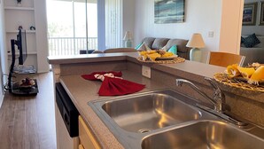 A modern kitchen with a large sink, a dining area with a sofa and a lamp, and a view of the outdoors through large windows. - Lovely Condo Stay Disney Orlando, 2Bed/2Bath (Kissimmee)