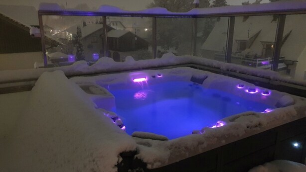 Vasca idromassaggio coperta di neve illuminata con luci viola, circondata da un recinto in vetro.