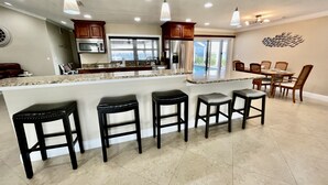 A modern kitchen with a central island, bar stools, a dining area with chairs, and a decorative wall piece. - Stunning open water view 3 bedroom house, pool, private dock (Cape Coral)