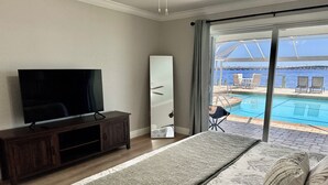 A bedroom with a large bed, a TV on a wooden cabinet, a mirror, and a view of an outdoor pool area. - Stunning open water view 3 bedroom house, pool, private dock (Cape Coral)