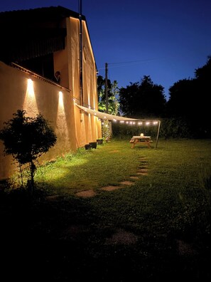 A building with a balcony, a string of lights, and a grassy area with a wooden swing set.