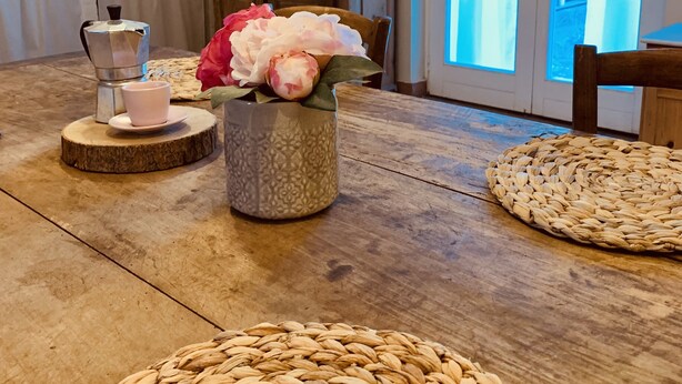 A kitchen with a wooden table, a coffee maker, and a vase with flowers. There are shelves with kitchen items and a window with curtains.