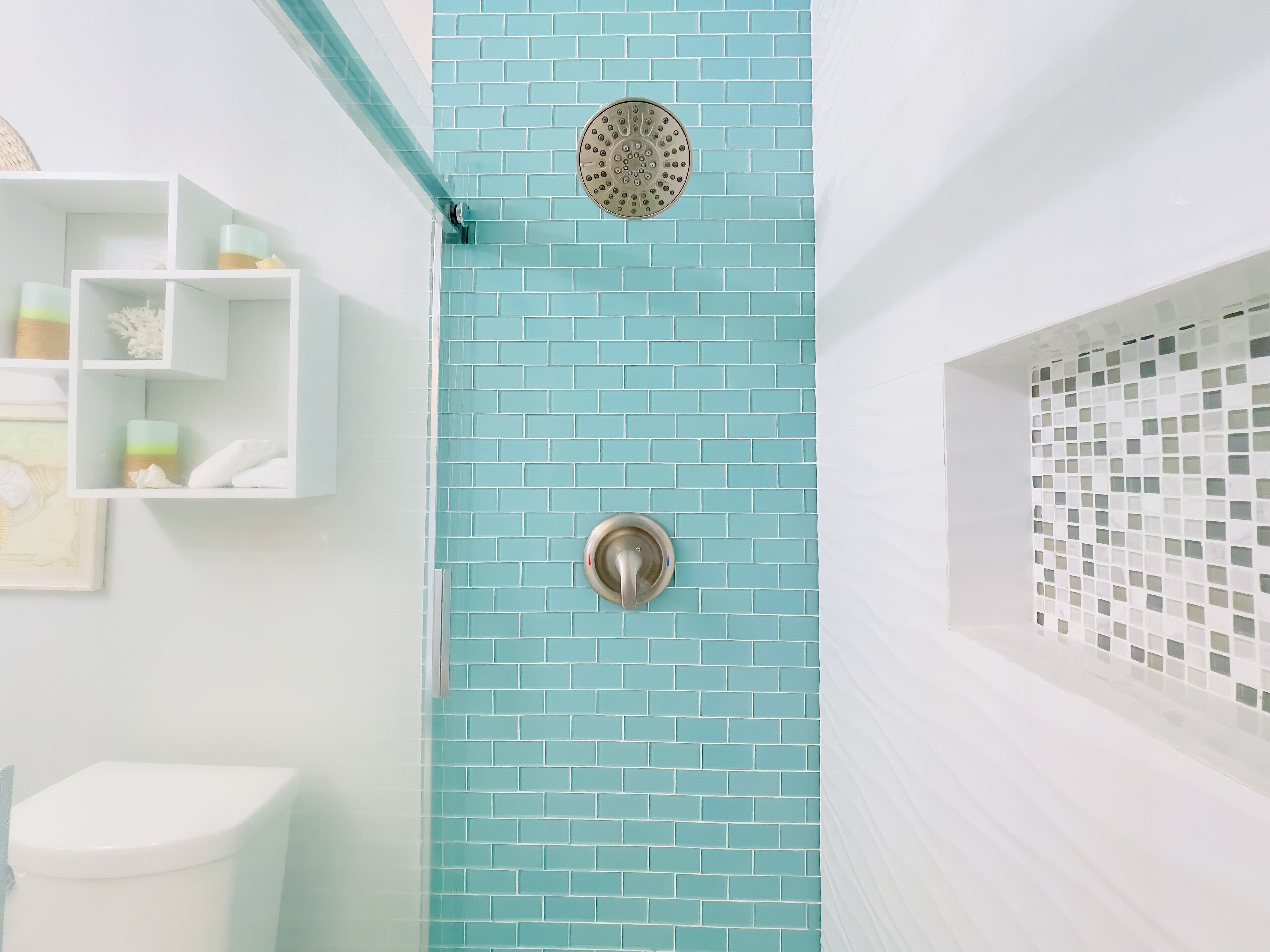 Une salle de bain avec un mur de douche recouvert de carreaux bleus, des toilettes blanches et une étagère contenant des articles de toilette.