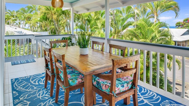 Een houten eettafel met bloemenkussens, een hangende hanglamp, en uitzicht op palmbomen en een huis door een witte balustrade.