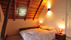 A cozy bedroom with a sloped ceiling, wooden beams, a large bed with a neatly folded blanket, and two bedside lamps.