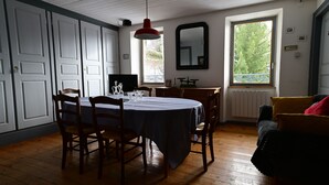 A dining area with a round table, chairs, a red pendant light, a window with a view of trees, and a radiator.
