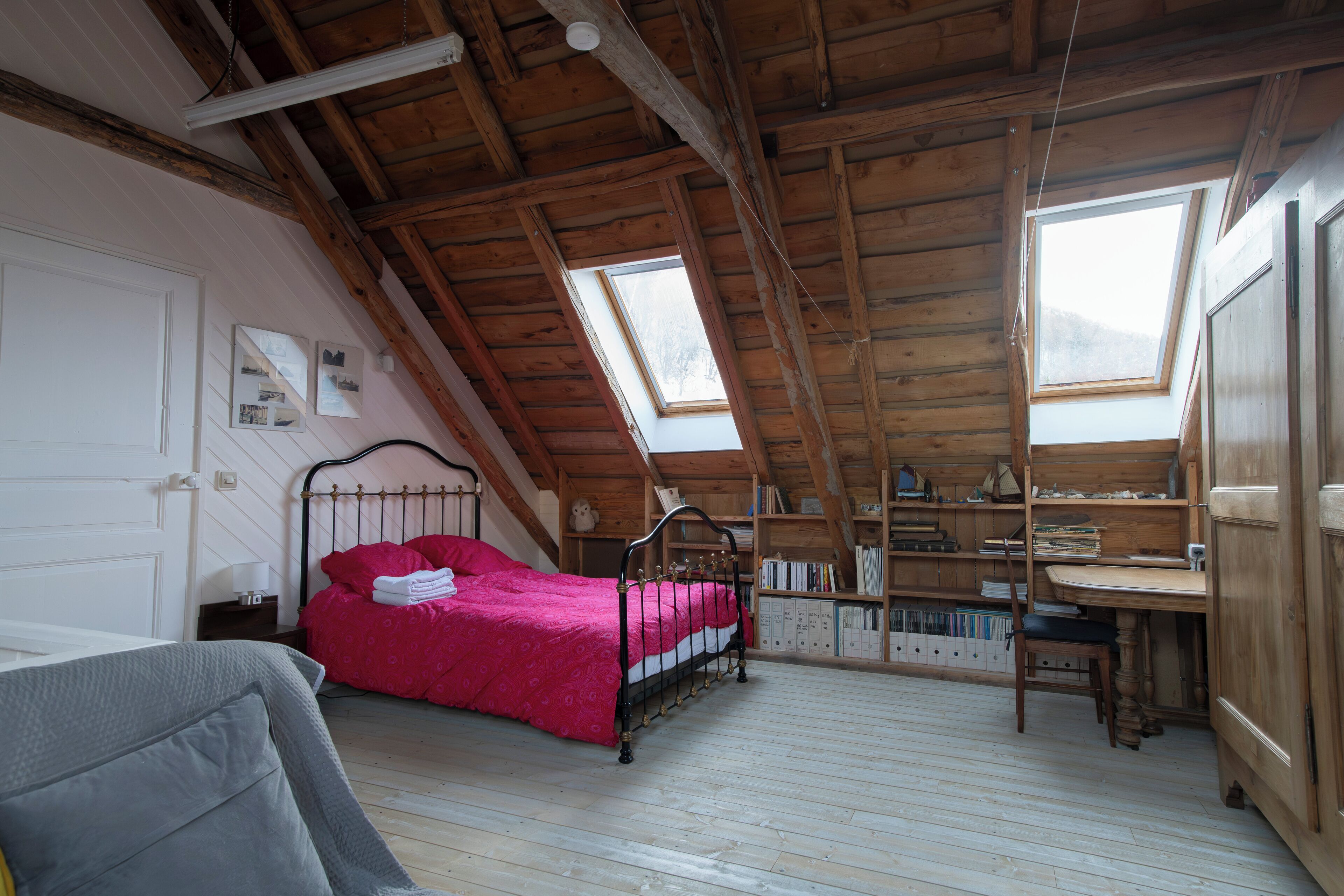 Une chambre avec un plafond en pente, un lit recouvert d’une couverture rouge, un bureau en bois et une bibliothèque.