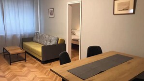Ett rum med ett trägolv i fiskbensmönster, en soffa med mönstrade kuddar, ett litet bord, ett träbord för matplatsen och två stolar.