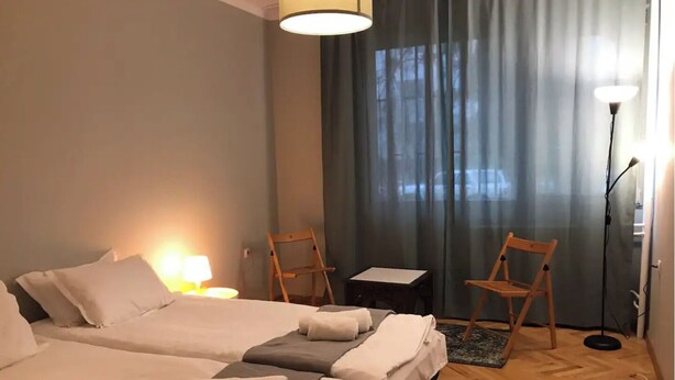 Ett hotellrum med två sängar, ett litet bord, två stolar, en lampa och utsikt över omgivningen genom ett fönster med gardiner.