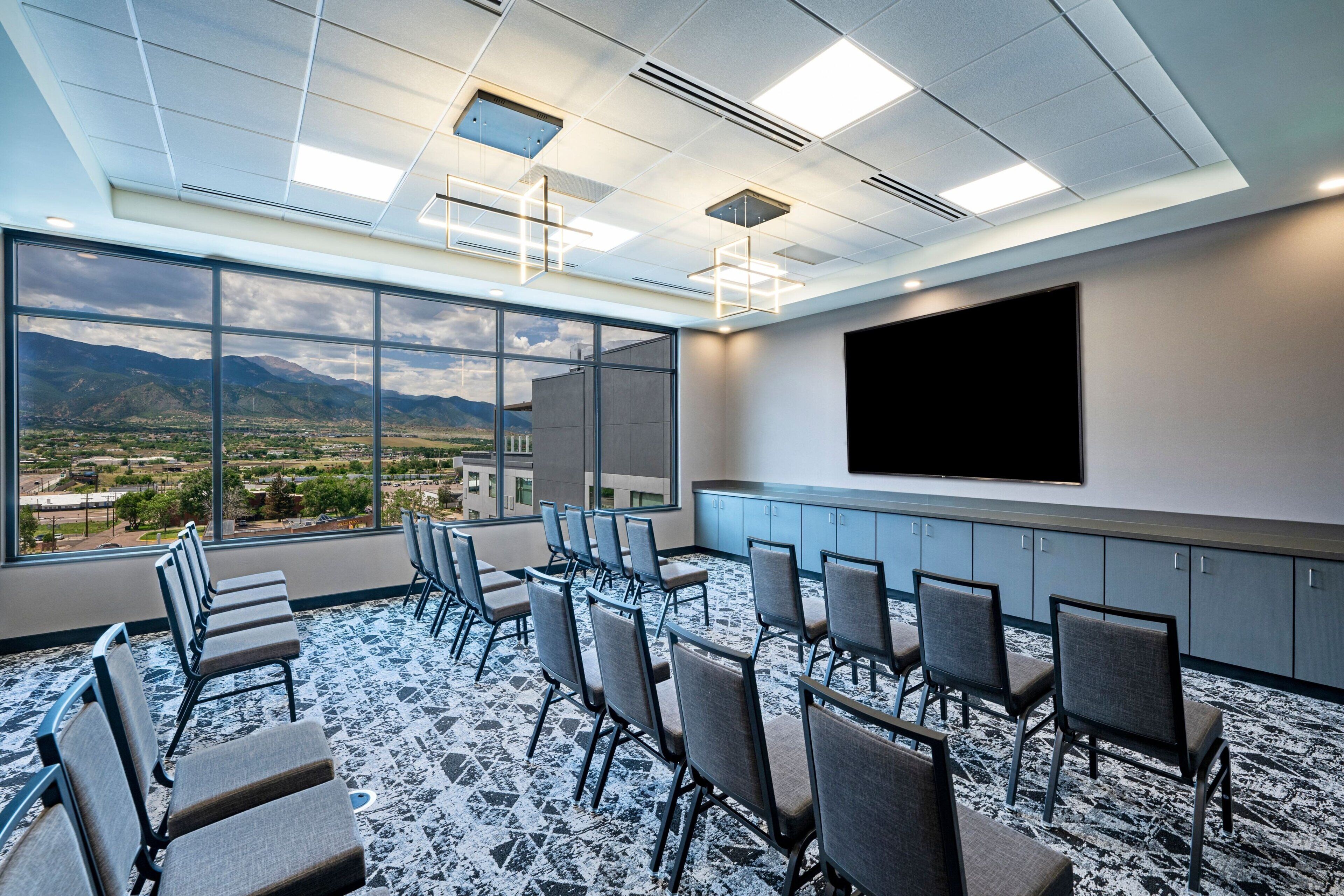 A conference room with a large window, chairs arranged in rows, a flat-screen TV, and a ceiling with recessed lighting.