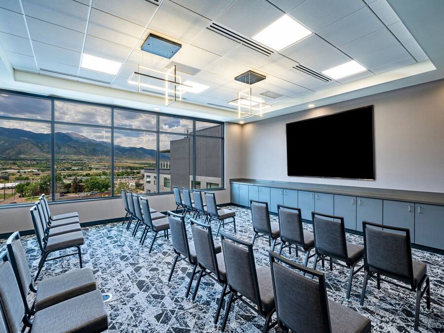 A conference room with a large window, chairs arranged in rows, a flat-screen TV, and a ceiling with recessed lighting.