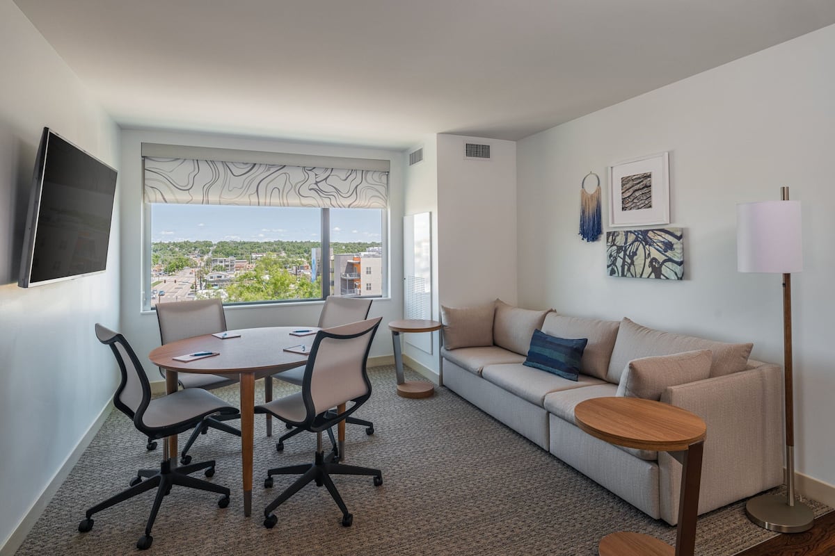 A modern hotel room with a large window offering a city view, a sofa, a dining table with chairs, and a TV mounted on the wall.