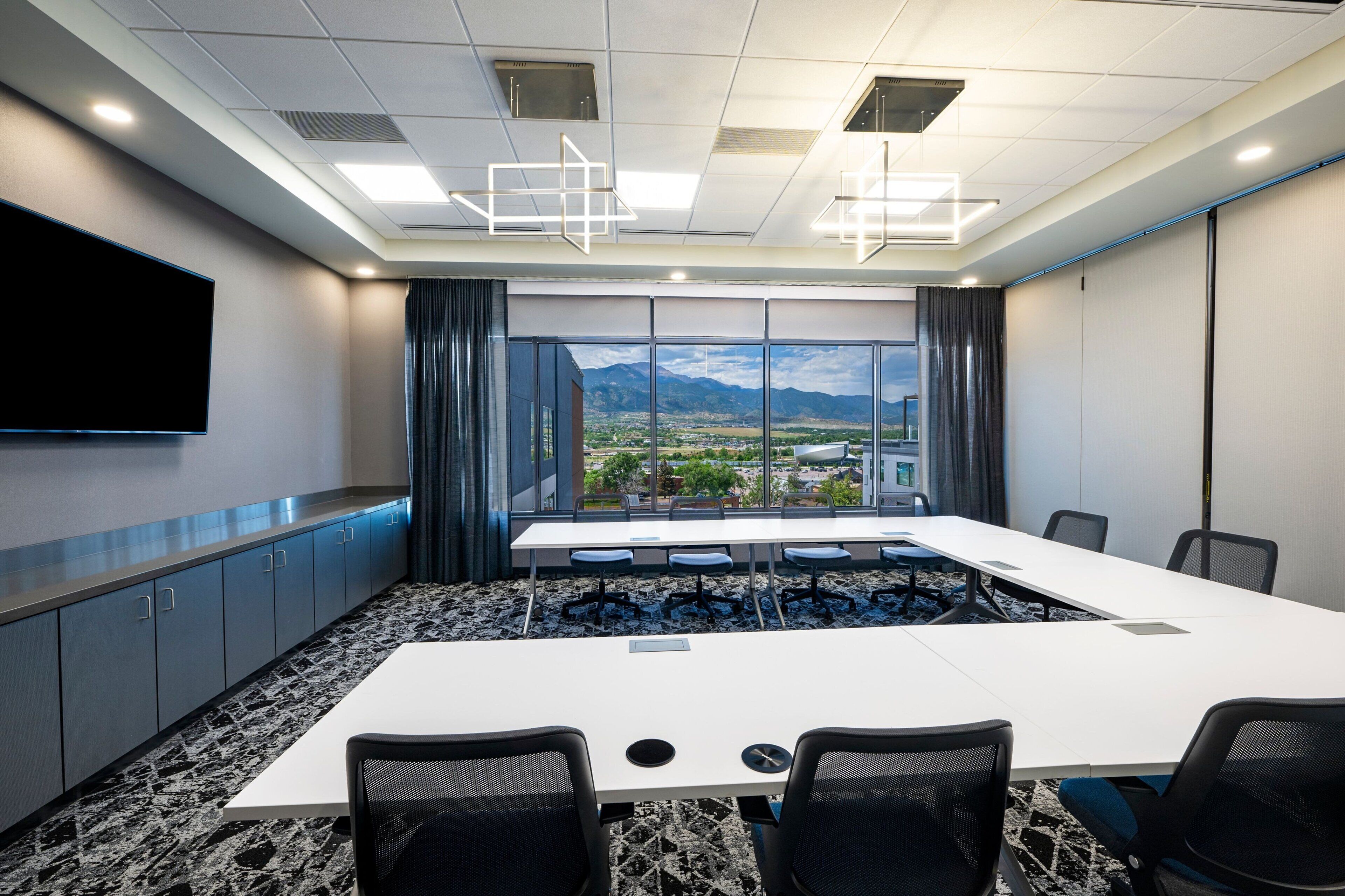 A conference room with a large table, chairs, a flat-screen TV, and a view of a cityscape through large windows.