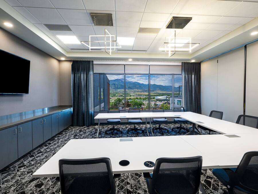 A conference room with a large table, chairs, a flat-screen TV, and a view of a cityscape through large windows.