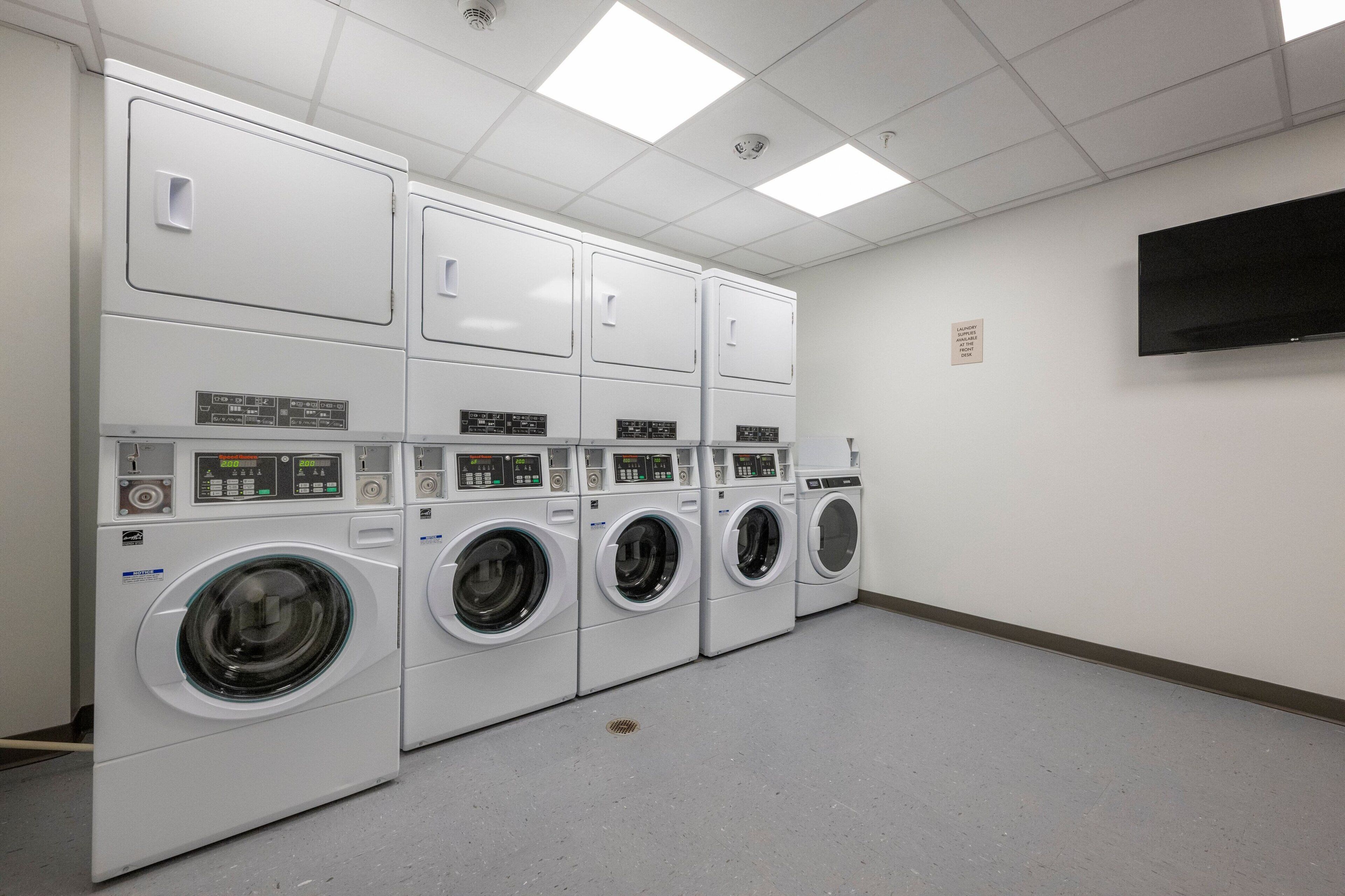 A laundry room with multiple washing machines and dryers, a flat-screen TV on the wall, and a tiled floor.