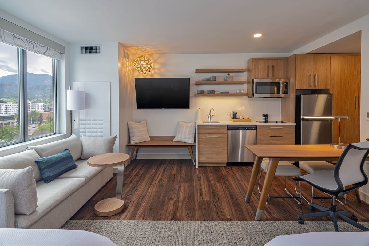 A modern hotel room with a sofa, a small round table, a dining table with chairs, a kitchen area with a microwave and refrigerator, and a large window offering a city view.