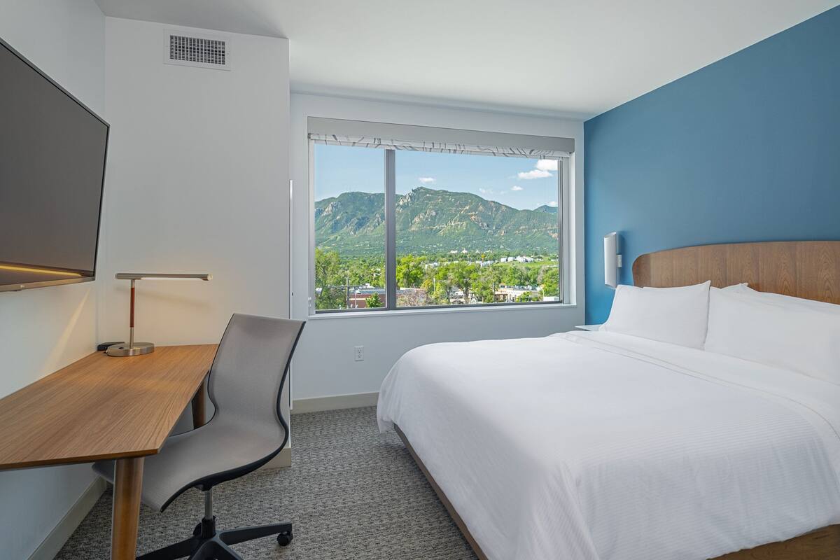 A hotel room with a bed, a desk, a chair, a TV, and a window with a mountain view.