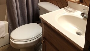 A bathroom with a wooden wall, a white toilet, a sink with a mirror, and a shower curtain. - Cabin #4   Secluded and Quiet (Oneida)