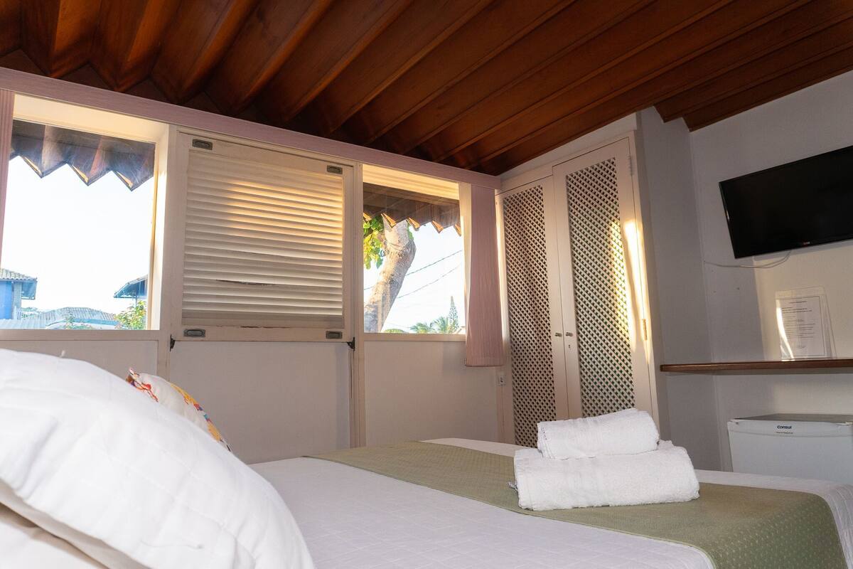 A hotel room with a bed, a TV, a window with blinds, and a view of the outside.