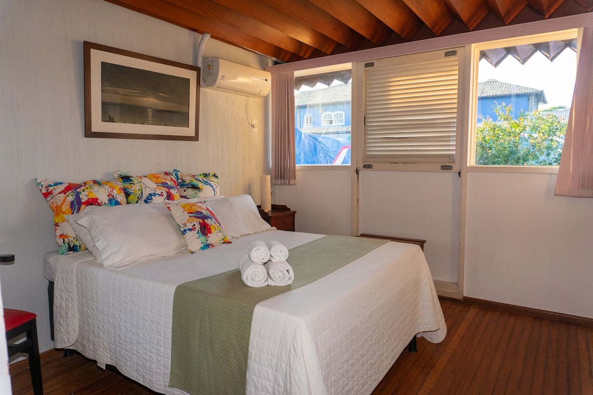 A neatly made bed with a white comforter and pillows, a green throw blanket, and rolled towels on the bed.
