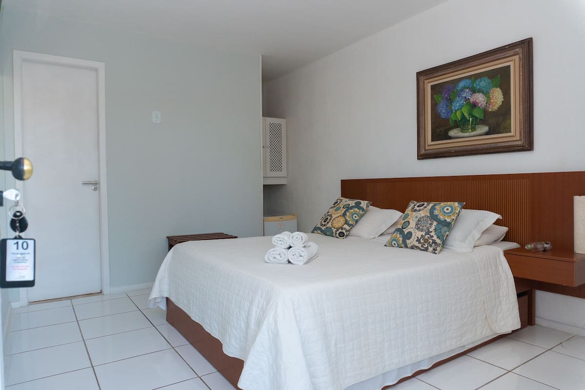 A neatly made bed with white bedding, a wooden headboard, and decorative pillows. A framed painting of flowers hangs on the wall above the bed.
