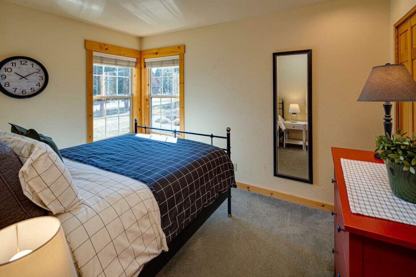 A bedroom with a bed, a bedside table with a lamp, a mirror, and windows with blinds.