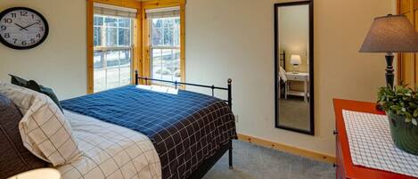 A bedroom with a bed, a bedside table with a lamp, a mirror, and windows with blinds.