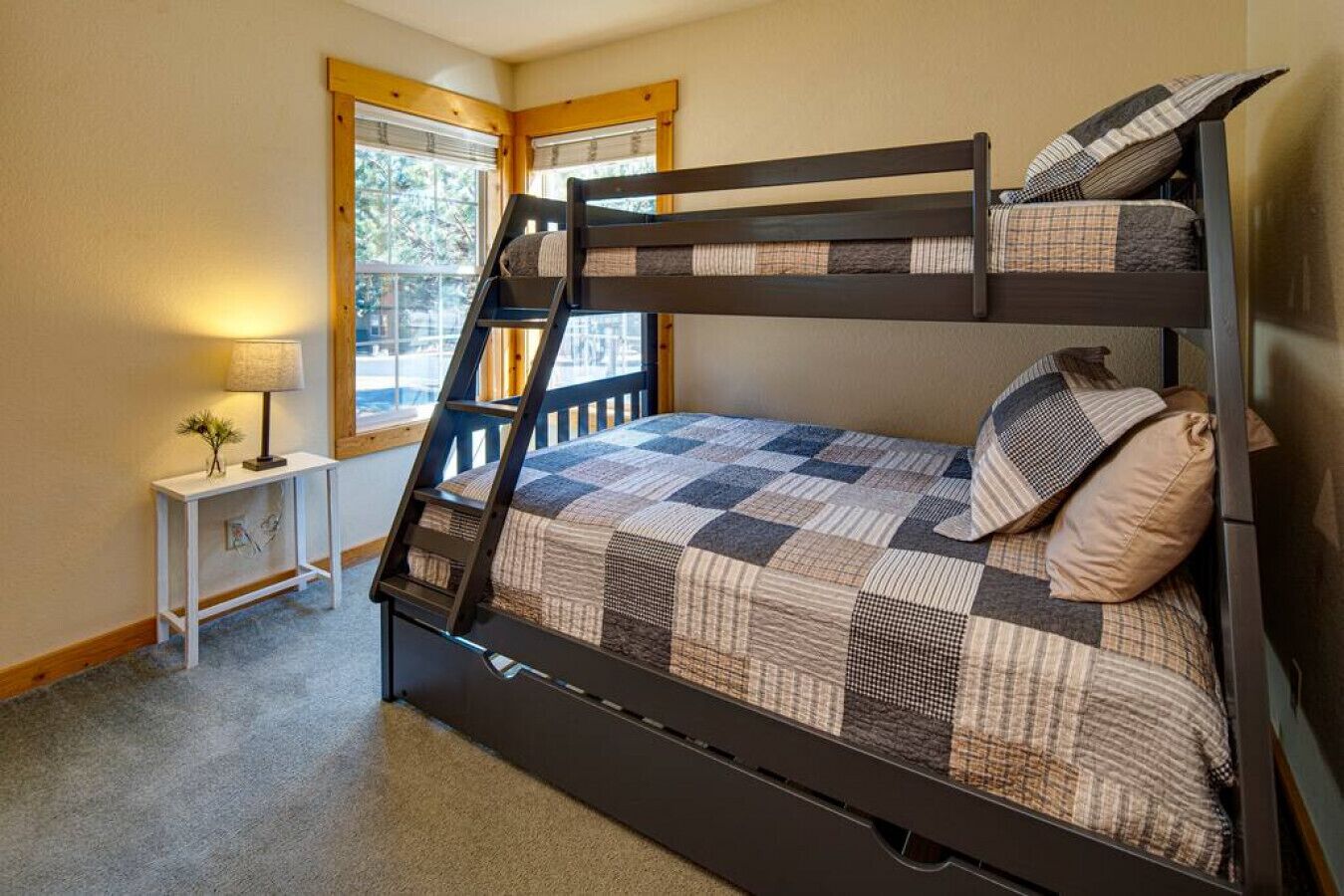 A bunk bed with a checkered bedspread and pillows, a nightstand with a lamp, and a window with blinds.