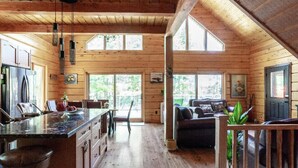 Interior de una cabaña con cocina, comedor y sala de estar, destacando paredes de madera y ventanas amplias.