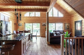 A cabin interior with a kitchen, dining area, and living room, featuring wooden walls and large windows.