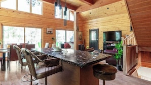 Interior de una cabaña de madera con comedor, un ventanal grande y una televisión.