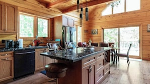 Cocina de cabaña de madera con una isla grande, armarios de madera y un comedor con vista al bosque.